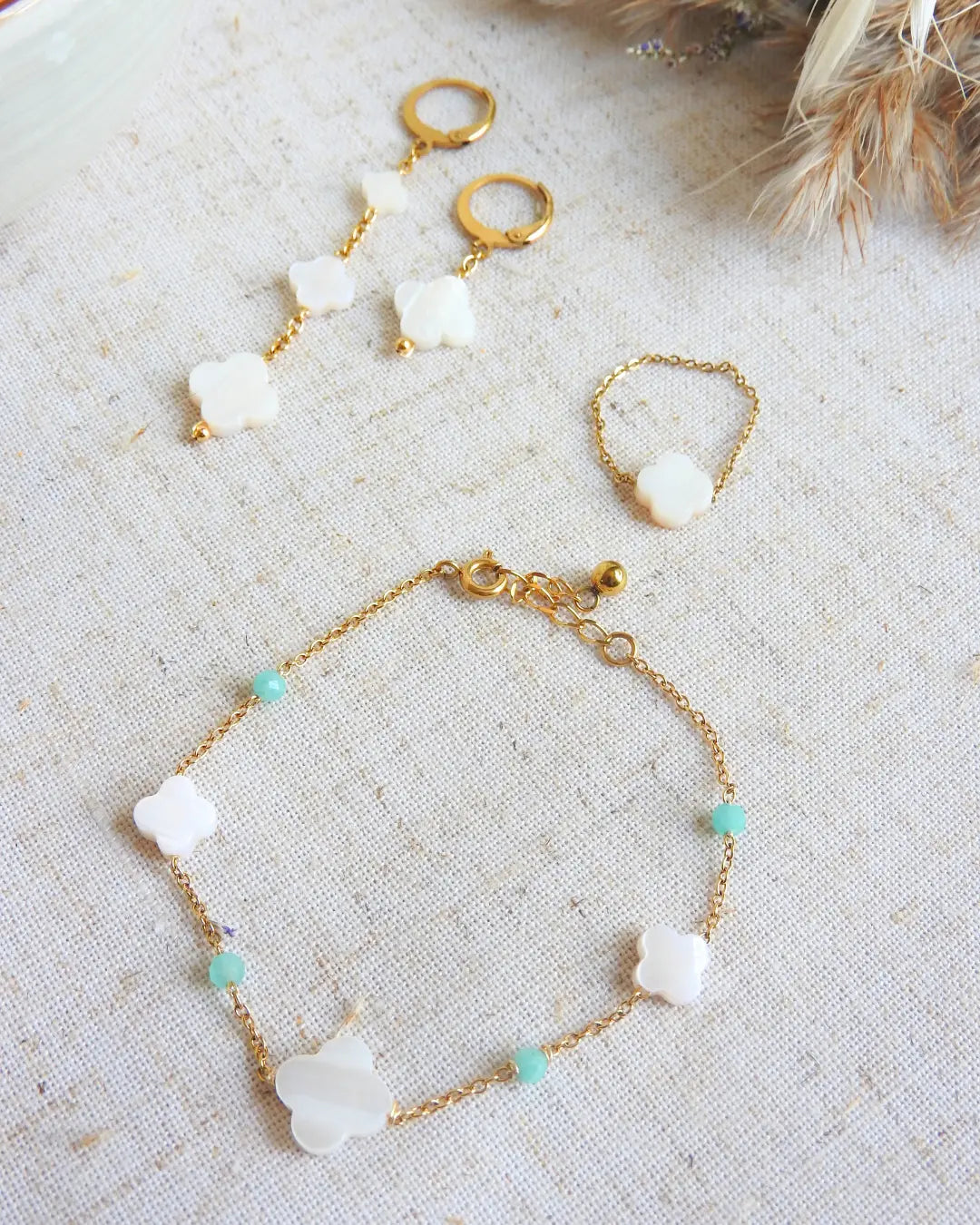 parure de bijoux posés à plat sur une nappe beige décorée de brindilles. Bracelet à chaîne perlée en perles de verre turquoise et perles trèfle en nacre blanche, une bague chaînette ornée d'une perle trèfle en nacre blanche et une paire de boucles d'oreilles à chainettes pendantes asymétriques avec des perles trèfle en nacre blanche.