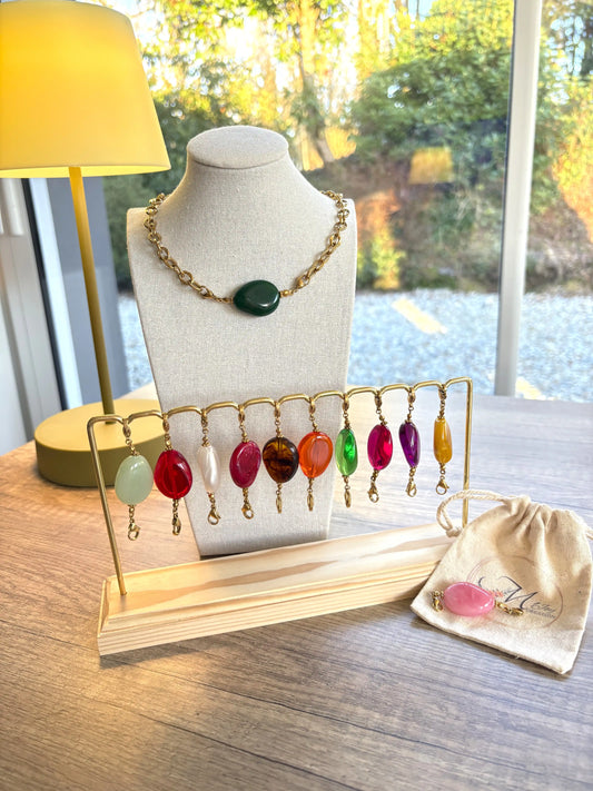 Perles colorées interchangeables de forme ovale irrégulière à positionner sur le collier majea. Perles alignées sur un présentoir a barre métalique et bambou. 