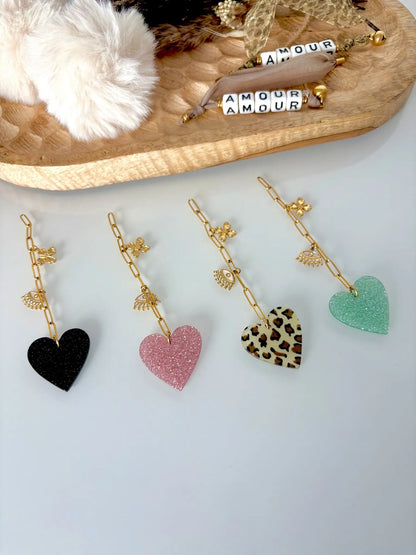 Chaînettes alignées les unes à côté des autres à breloques en acier inoxydable doré : trèfle, oeil protecteur et coeur paillette noir, rose, vert et motif léopart. Bijoux posés à plat sur une table en verre décorée d'un plateau en bambou avec des pompons beige.