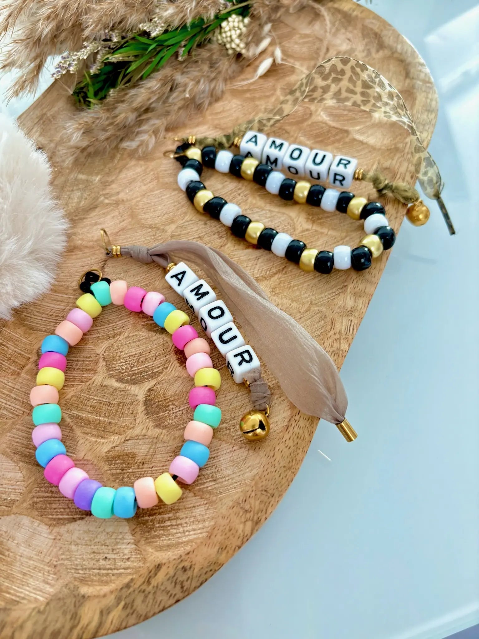 deux portes-clés alignés l'un à côté de l'autre. Bracelets à perles multicouleurs pour l'un avec un ruban moka orné de perles cubes blancs formant l'inscription AMOUR et finissant avec un grolet en acier inoxydable doré. L'autre avec un bracelet de couleurs noir, blanc et doré avec un ruban au motif léopard avec l'inscription AMOUR en perles cubes blancs finissant avec un grelot doré.