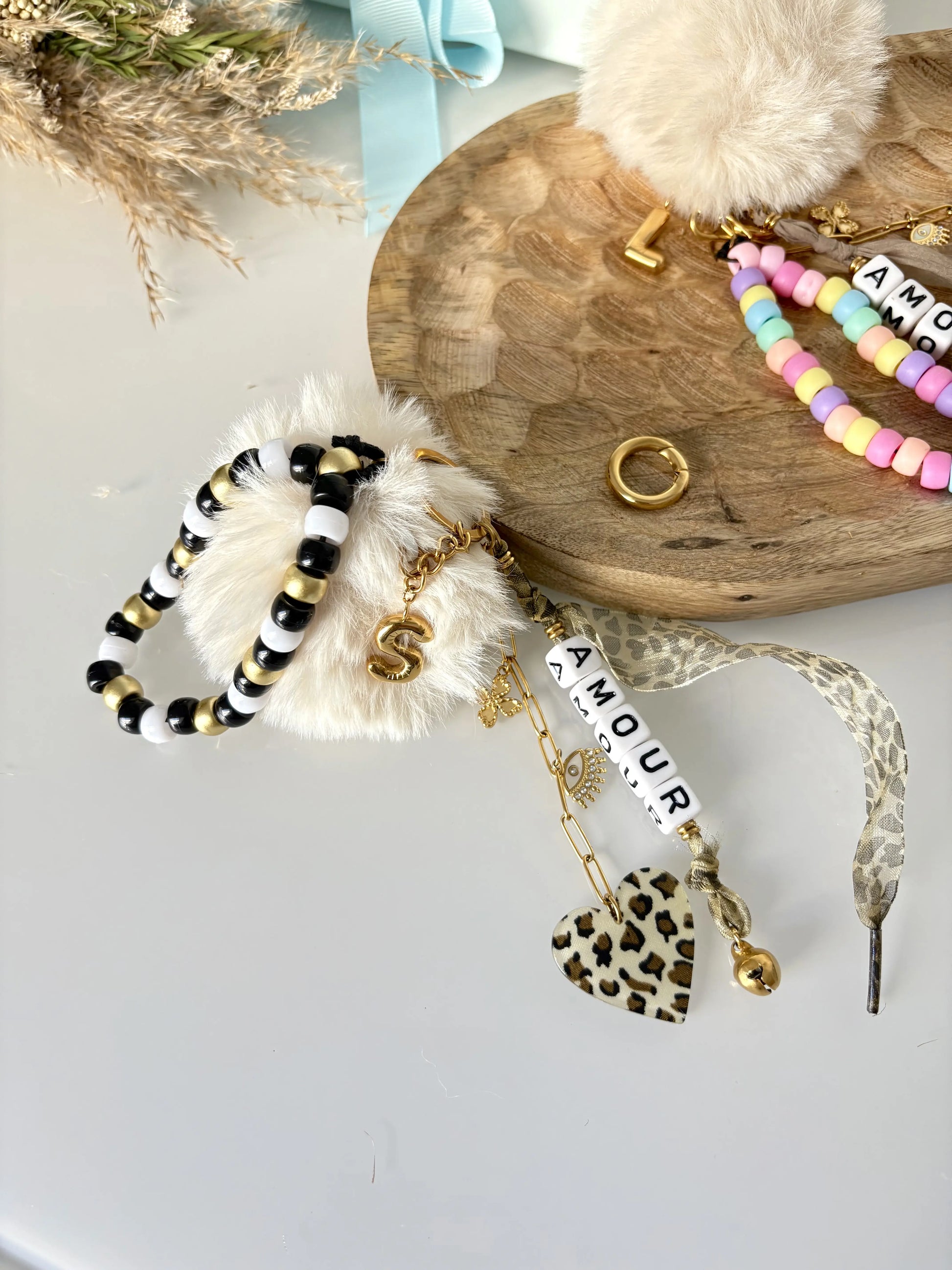 deux portes-clés ou bijoux de sac posés à plat sur une table et un plateau en bambou. Bijoux décorés d'un bracelet de perles de couleurs noir, blanc et doré, un ruban et un coeur au motif léopard, une lettre S à effet ballon en acier inoxydable doré et une chaînette doré avec des breloques trèfle et oeil protecteur.