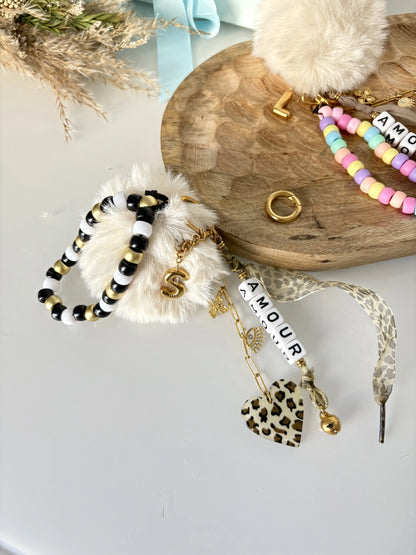 deux portes-clés ou bijoux de sac posés à plat sur une table et un plateau en bambou. Bijoux décorés d'un bracelet de perles de couleurs noir, blanc et doré, un ruban et un coeur au motif léopard, une lettre S à effet ballon en acier inoxydable doré et une chaînette doré avec des breloques trèfle et oeil protecteur.