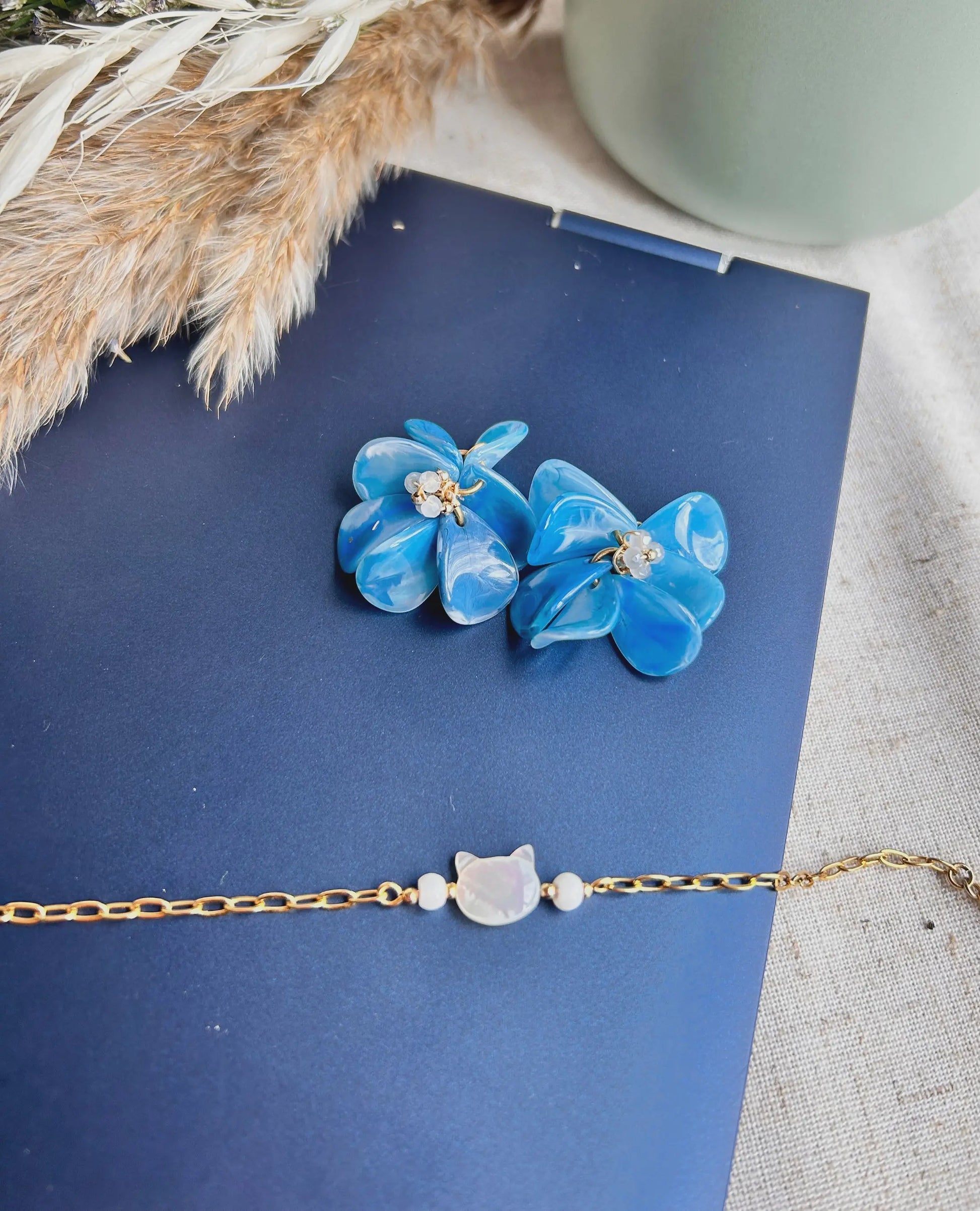 Suspensions pour créoles en acier inoxydable assemblées à la main. Bouquet floral réalisé à partir de pétales en acrylique bleu marbré et de perles de verre colorées et en acier inoxydable doré. Bijou posé sur le dessus d'un ordinateur portable avec un bracelet chaîne maille rectangle en acier inoxydable doré, décoré d'une perle chat en nacre blanche. 